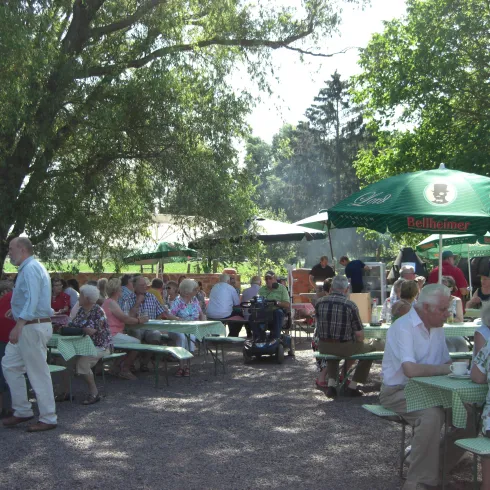 Eine gesellige Versammlung von Menschen in einem Biergarten. Tische sind mit grünen Tischdecken dekoriert, und Sonnenschirme spenden Schatten.