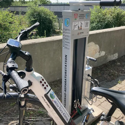 Eine Fahrradreparaturstation steht neben einem Fahrrad. Im Hintergrund sind Bäume und ein blauer Himmel zu sehen.