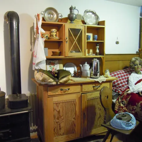 Ein gemütliches Zimmer mit einem Holzschrank und historischen Küchenutensilien. Auf dem Bett sitzt eine lebensechte Puppe, die die Atmosphäre vergangener Zeiten widerspiegelt.