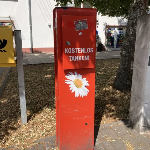 Eine rote Säule mit der Aufschrift "KOSTENLOS TANKEN!" und einer weißen Blume. Sie steht auf einem Gehweg im Freien, umgeben von Bäumen.