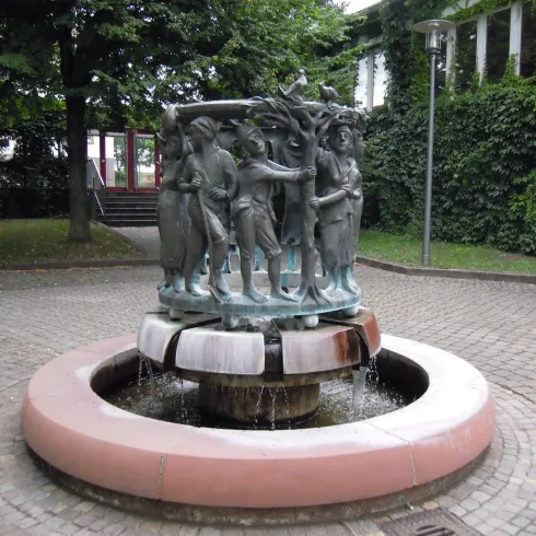 Ein kunstvoller Brunnen mit einer Skulpturengruppe aus Bronze steht in einem Garten. Umgeben von Bäumen und Sträuchern sprudelt das Wasser sanft.