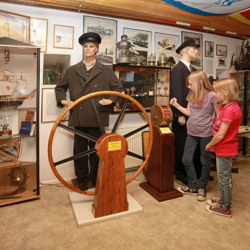 Im Bild sind zwei Kinder zu sehen, die ein Steuerrad in einem Museum betrachten. Neben ihnen stehen zwei Schaufensterpuppen in maritimer Kleidung.