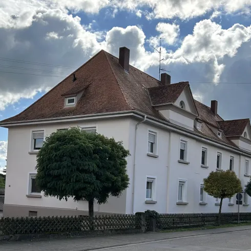 Ein großes, weißes Gebäude mit einem roten Dach und großen Fenstern. Mehrere Bäume stehen im Vordergrund vor einem bewölkten Himmel.