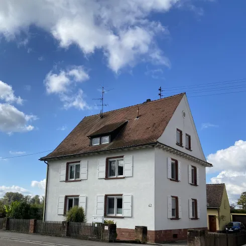Ein weißes Haus mit einem braunen Dach und mehreren Fenstern. Der Himmel ist blau und mit einigen Wolken bedeckt.