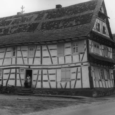 Ein historisches Fachwerkhaus mit einem schrägen Dach und traditioneller Holzverzierung. Es steht in einer ländlichen Umgebung und zeigt die Architektur vergangener Zeiten.