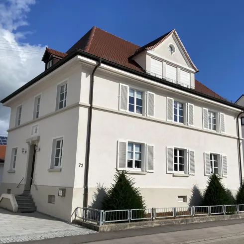 Ein modernes, zweistöckiges Haus mit weißen Wänden und roten Dachziegeln. Vor dem Haus stehen einige kleine immergrüne Bäume.