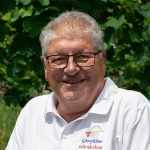 Ein lächelnder Mann mit Brille trägt ein weißes T-Shirt und sitzt vor üppigem Grün. Im Hintergrund sind Weinreben zu sehen.